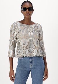 Beige Spitze-Oberteil mit aufwendiger floralem Stickerei und Dreiviertelärmeln, kombiniert mit hautengen, hochgeschnittenen blauen Jeans. Schwarze Sonnenbrille getragen.
