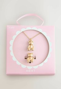 Guldtonet bjørnecharms halskæde og matchende studs øreringe i en cirkulær udstillingspakke med pink base og bølgede kanter.