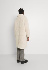 Abrigo de piel sintética color crema con cuello alto, mangas largas y corte recto, llevado sobre pantalones de cuadros, sobre un fondo blanco liso.
