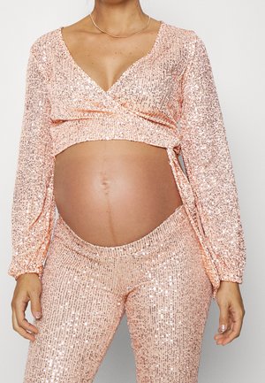 Personne enceinte portant un haut cache-cœur en sequins rose clair étincelant et un pantalon assorti, montrant le ventre nu et les mains sur les côtés.