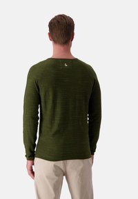 Grünes Langarmshirt mit strukturierter Oberfläche, Rundhalsausschnitt und einem kleinen Etikett mit einem kaufmännischen Und-Zeichen auf der Rückseite.