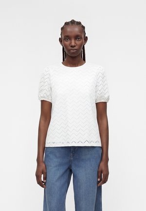 Γυναίκα με πλεξούδες, που φοράει λευκό κοντομάνικο μπλουζάκι με ζιγκ-ζαγκ μοτίβο με οπές και μπλε τζιν, όρθια μπροστά σε μονόχρωμο φόντο.