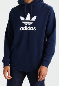 Person som bär en marinblå Adidas-hoodie med vitt treklöverlogo och text, tillsammans med mörkblå jeans.