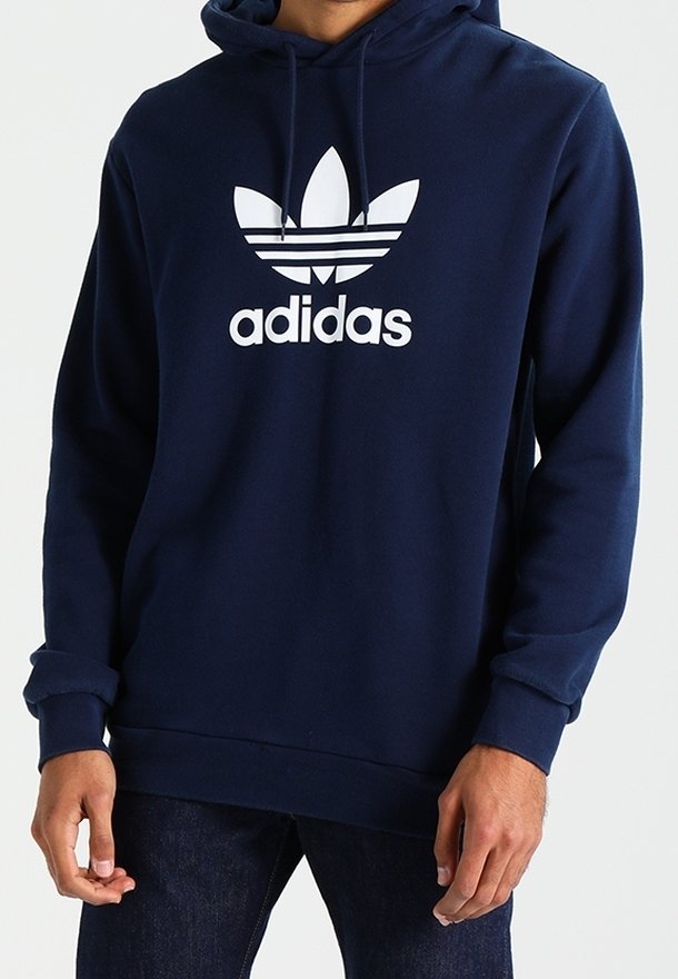 Person som bär en marinblå Adidas-hoodie med vitt treklöverlogo och text, tillsammans med mörkblå jeans.