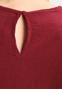 Burgundy top med design med nøglehul i ryggen, fremstillet af et tekstureret stof med subtile stikninger langs halsudskæringen og kanterne.