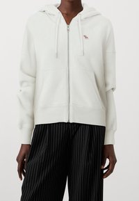 White zip-up hoodie with small red moose logo on chest, worn with black pinstripe wide-leg trousers by a standing person.