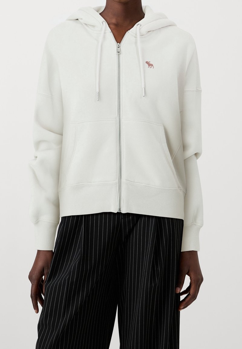 White zip-up hoodie with small red moose logo on chest, worn with black pinstripe wide-leg trousers by a standing person.