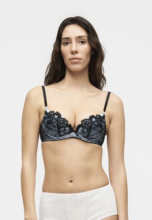 Kobieta z długimi ciemnymi włosami, ubraną w czarny koronkowy biustonosz i białe majtki z wysokim stanem, stojąca na jednolitym jasnym tle.