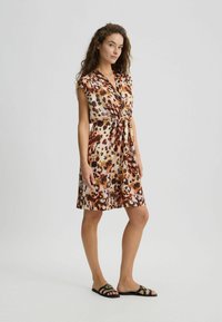 Abito con stampa animale in tessuto leggero, con scollo a V, maniche corte e vita con cintura. I colori includono crema, marrone e ruggine.