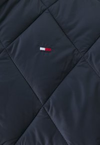 Mörkt quiltat tyg med diamantstygn och en liten broderad röd och vit rektangulär logotyp i mitten.