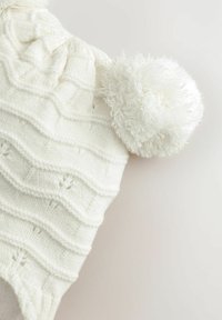 Hvid strikket hat med et tekstureret bølgemønster, der har en fluffy pom-pom på toppen. Materialet ser blødt og hyggeligt ud.