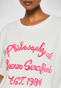 Close-up of person wearing white shirt with bright pink script text "Philosophy di Lorenzo Serafini EST. 1984" on front.