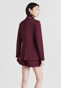 Blazer bordeaux con una vestibilità su misura, caratterizzato da un colletto a punta e una fessura posteriore singola, abbinato a pantaloni corti coordinati. Tessuto liscio.