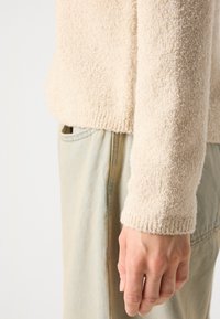 Krém színű, texturált pulóver, bordás mandzsettákkal, világoskék farmernadrággal párosítva, közeli felvétel a kar és kéz részéről.