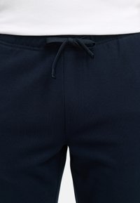 Gros plan sur un pantalon bleu marine avec une taille à cordon noué, porté avec une chemise blanche rentrée à la taille.