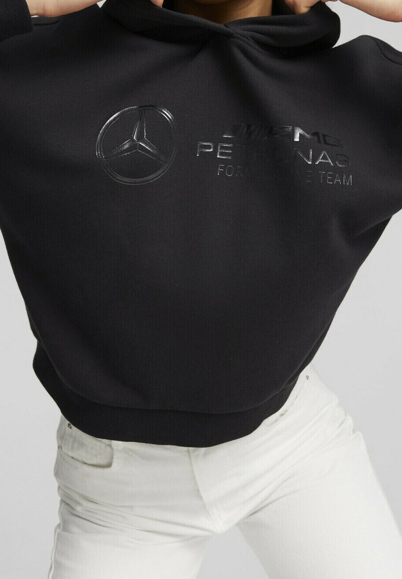 Černá zkrácená mikina s kapucí vyrobená z texturované látky, s hladkým reliéfním logem Mercedes a nápisem "PETRONAS FORMULA ONE TEAM" v tučném písmu.
