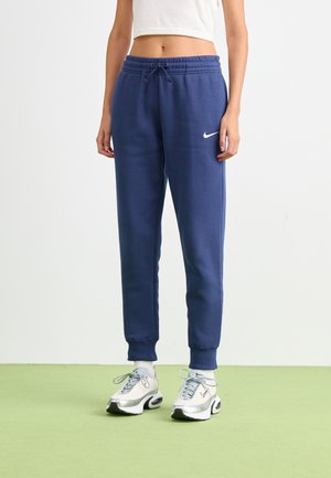 Persoon in marineblauwe Nike-sweatpants, wit cropped top, witte sokken en witte en zilveren Nike sneakers staat op een lichtgroene vloer.