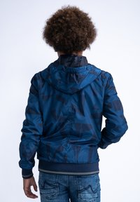 Kapsáčová bomber bunda s kapucí v tmavě modré barvě s geometrickým vzorem, lehký materiál, elastické manžety a stáhnutý pas.