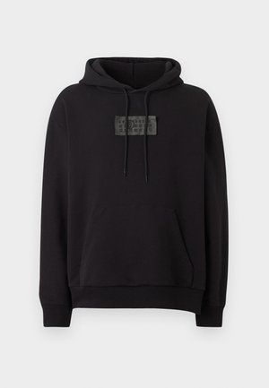 Giovane uomo con felpa nera con cappuccio e logo frontale, pantaloncini neri al ginocchio, calzini bianchi e mocassini neri, in piedi su sfondo bianco.