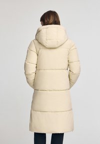 Cappotto imbottito beige con cappuccio, caratterizzato da un design trapuntato e una silhouette lunga, texture liscia e maniche aderenti.