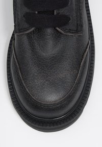 Przednia część zużytego czarnego skórzanego buta z widocznymi zagnieceniami, grubą, żłobioną podeszwą i czarnymi sznurówkami na białym tle.