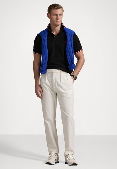 Homem a usar uma camisola polo preta, calças brancas plissadas, sapatilhas bege e um casaco azul a pender nos ombros, de pé com as mãos parcialmente nos bolsos.