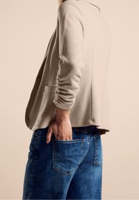 Beige pletený blazer s dlouhými rukávy a knoflíkovými detaily, spárovaný s modrými džíny s zadními kapsami a jemně ošetřenou texturou.