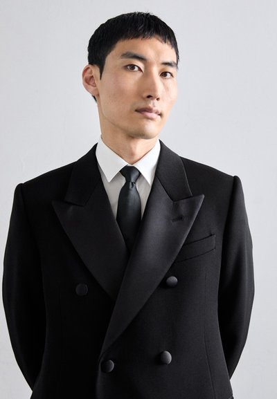 Melna dubultā pakalpojuma uzvalka jaka ar satīna apmalēm, saskaņota ar baltu svinīgu kreklu un melnu kaklasaiti. Gluda auduma, pielāgota piegriezuma, minimālisma stila.