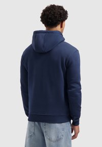 Man met krullend haar in een effen marineblauwe hoodie en lichte spijkerbroek, met zijn rug naar de kijker op een lichtgrijze achtergrond.