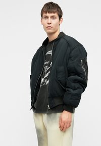 Fekete bomber dzseki cipzáros elülső résszel, bordás mandzsettákkal és egy oldalsó zsebbel; egy szürke mintás pólóval és világos színű nadrággal viselve.