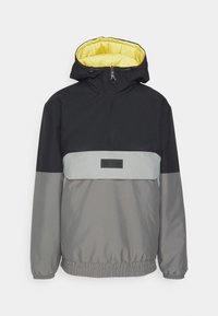 Chaqueta tipo pullover con una parte superior negra, una sección inferior gris y una banda plateada en el pecho. Forrada de amarillo y con puños elásticos.