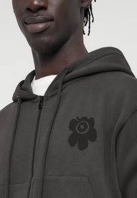 Σκούρα γκρι φούτερ με φερμουάρ, με μεγάλο μαύρο λουλουδάτο σχέδιο στην αριστερή πλευρά του στήθους και τσέπη καγκουρό μπροστά.