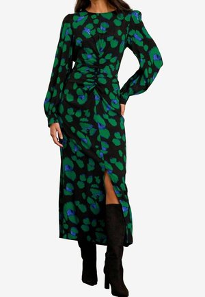 Vrouw draagt een zwarte jurk met lange mouwen met groene en blauwe abstracte vlekken, voorsplit, gecombineerd met zwarte kniehoge laarzen.