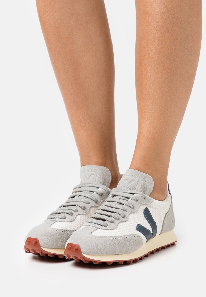 Sapatilhas cinza claro e brancas com um cabedal em malha, detalhes em camurça e acabamentos em azul escuro. Apresenta uma sola de borracha castanha com aderência texturizada.