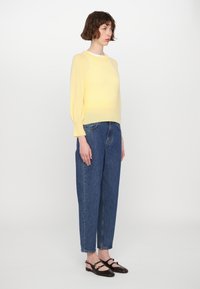 Gelber Strickpullover mit langen Puffärmeln, kombiniert mit hochgeschnittenen blauen Jeans. Braune Slipper mit Schnallen.