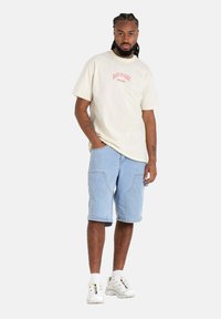 Beige katoenen t-shirt met rode tekst, gecombineerd met lichtblauwe denimshorts. Witte sneakers met accenten. Glad materiaal, casual pasvorm.