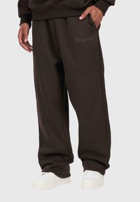 Person wearing loose dark brown sweatpants with "Dropsie" logo on the thigh and white sneakers, hand in pocket against plain background.