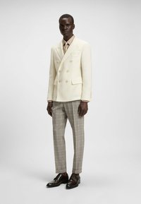 Blazer avorio doppiopetto con bottoni dorati, camicia beige e pantaloni a quadri nei toni di grigio e marrone; scarpe in pelle marrone.