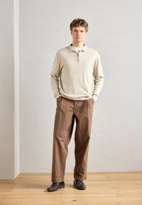 Pull en tricot beige avec fermeture éclair à l'avant, associé à un pantalon large marron. Des chaussures noires complètent l'ensemble, le tout sur un fond neutre.