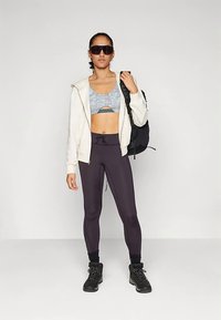 Sportos öltözék világosszürke mintás sportmelltartóval, krémszínű kapucnis pulóverrel, fekete leggingsszel és fekete csizmával. Egy hátizsák az egyik vállra akasztva.