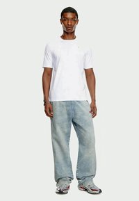 Young man standing wearing a plain white t-shirt, light blue baggy jeans, silver sneakers, and a bracelet on left wrist.