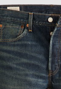 Gros plan sur un jean en denim bleu foncé montrant les poches avant, les rivets en cuivre, la passants de ceinture et le bouton métallique avec des coutures visibles.