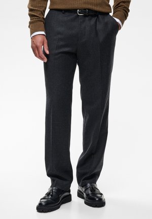 Mann trägt graue maßgeschneiderte Hose, schwarzen geflochtenen Gürtel, braunen Pullover und schwarze Lederloafer mit Quasten, steht mit Händen in den Taschen.