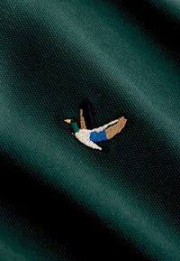 Tela verde con detallado bordado de un pato azul en vuelo, con pico amarillo, cuello blanco, parche azul en el ala y plumas marrones en la cola.