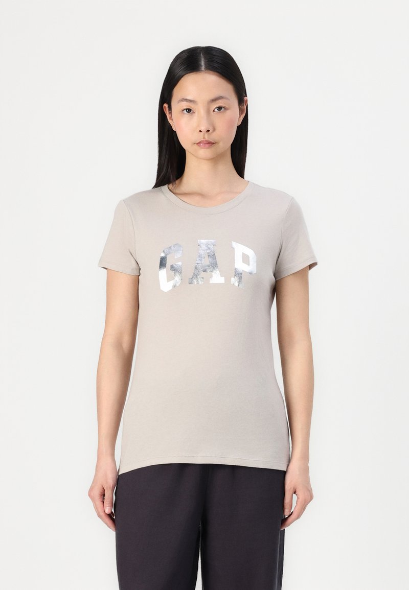Beige bomulls t-skjorte med kort erm, rund hals og sølvtrykt "GAP"-logo. Stoffet ser mykt og glatt ut.
