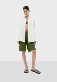 Bjela oversized košulja s dugim rukavima, zelene kratke hlače, crne japanke, model stoji na bijeloj pozadini, prikazujući ležeran, opušteni stil.