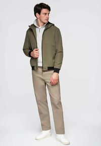 Zielona oliwkowa kurtka bomberka z kapturem, czarnymi mankietami i kieszeniami z przodu, noszona na jasnoszarym hoodie; zestawiona z beżowymi spodniami i białymi trampkami.