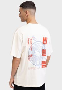 T-shirt in cotone color crema con grafica sul retro di spaghetti e testo in rosso e blu. Maniche corte, vestibilità ampia e senza loghi visibili.