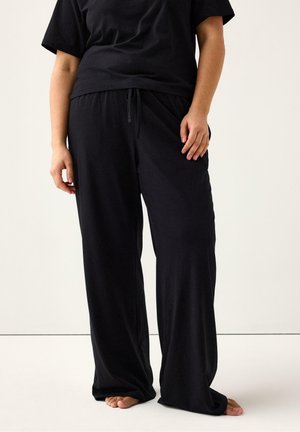 Persoon draagt losse zwarte broek en een zwart overhemd, staat blootsvoets op een effen witte vloer met een neutrale achtergrond.