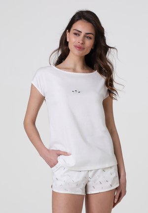 Donna con capelli lunghi e scuri che indossa una maglietta bianca a maniche corte e pantaloncini bianchi con piccoli motivi di foglie scure, in piedi con una mano in tasca.